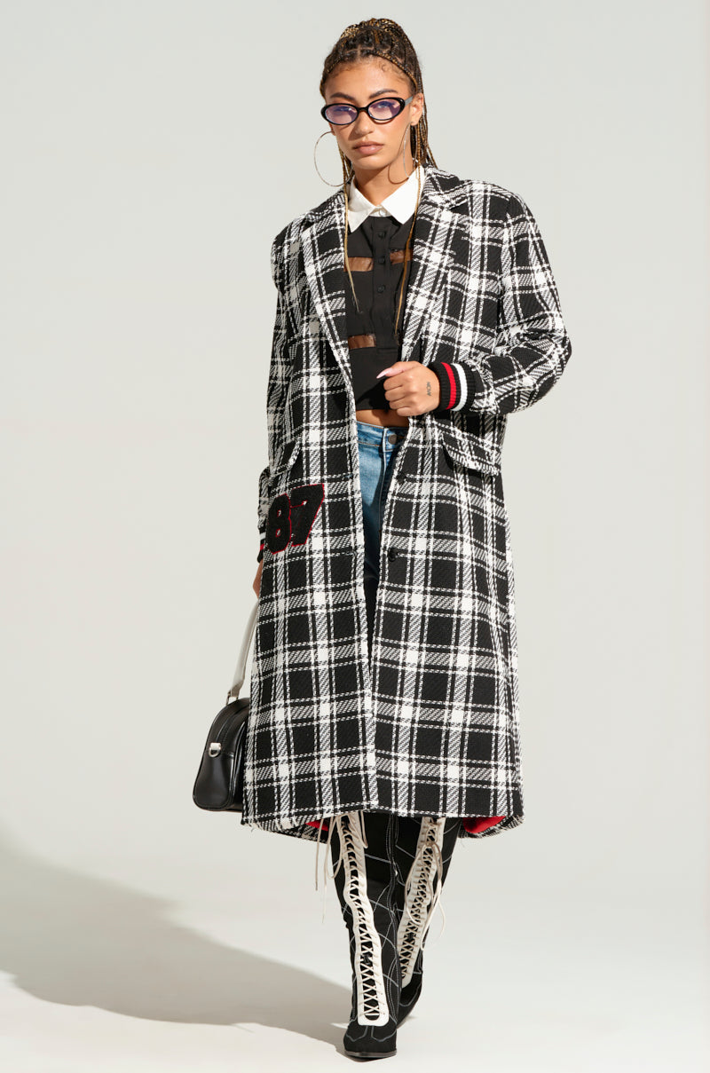 A person is wearing a long black and white plaid LIBERTY VARSITY TRENCH, a black blouse, blue jeans, and white lace-up boots. They have braided hair, hoop earrings, and purple glasses, holding a black handbag while walking confidently on a light-colored background.