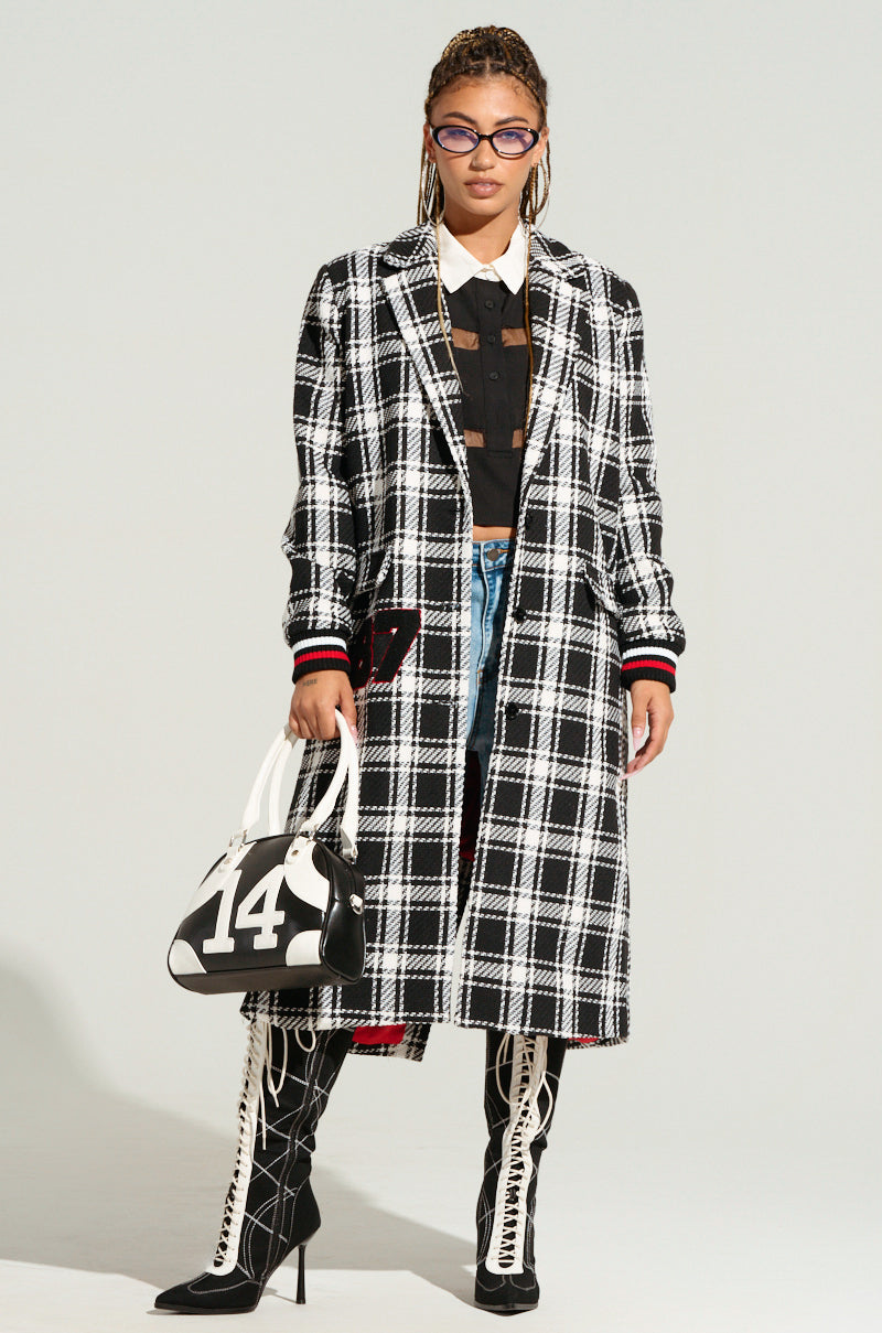 A woman with braided hair is sporting purple sunglasses, a black and white plaid LIBERTY VARSITY TRENCH, and knee-high black boots with white laces. Carrying a black and white handbag adorned with the number "14" and letterman-inspired patches, she stands against a plain background.