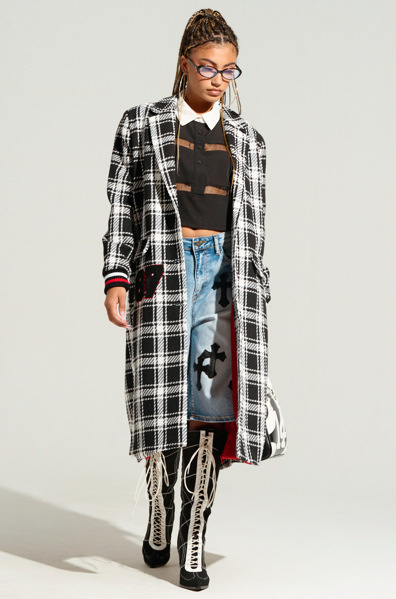 A person with braided hair wearing glasses walks down a runway in the LIBERTY VARSITY TRENCH adorned with letterman-inspired patches. They sport a black crop top, a blue denim skirt featuring graphic prints, and white high-laced boots. Complementing their look is a matching black-and-white bag.