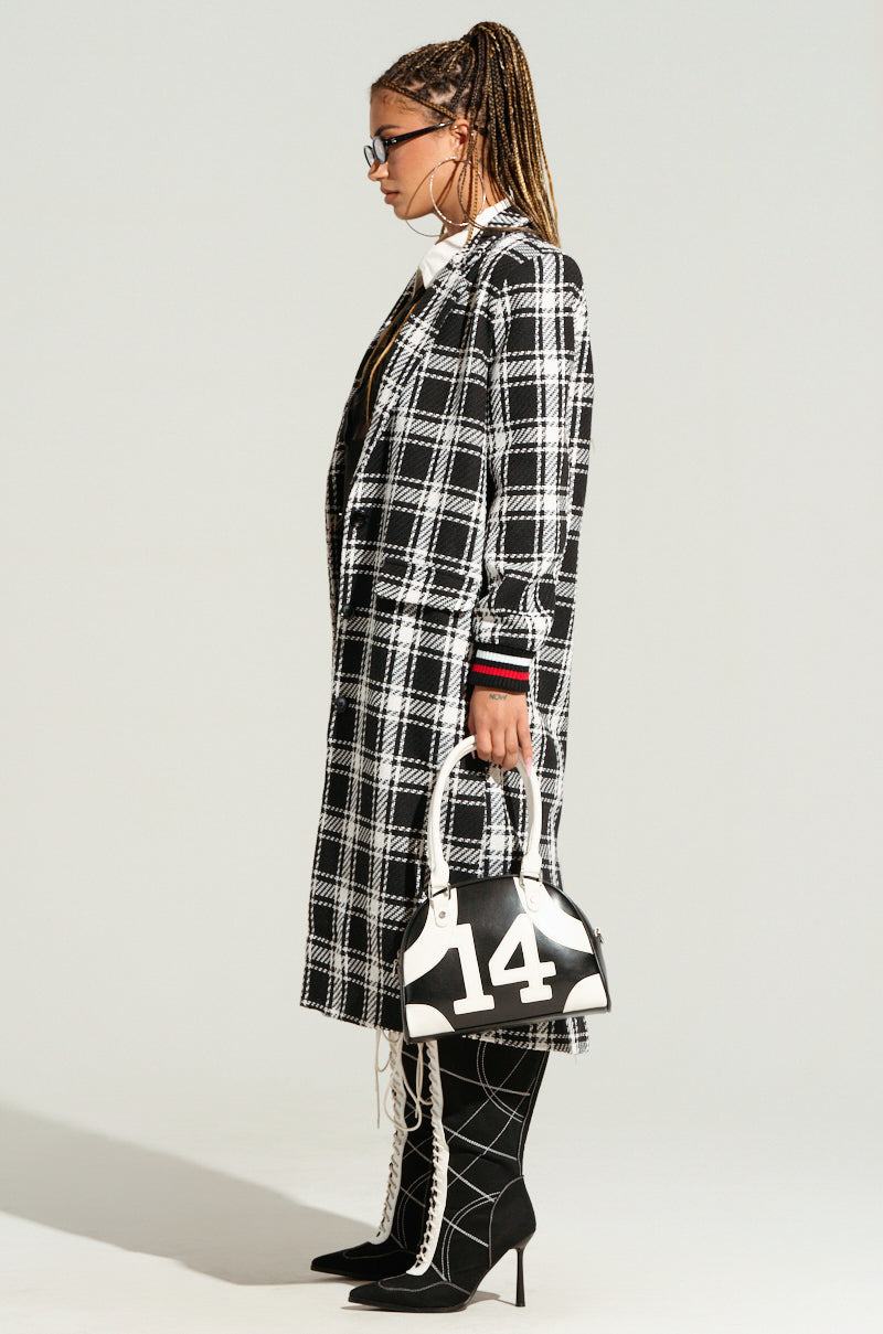 A person with long braided hair stands in profile against a plain background, wearing a black and white plaid LIBERTY VARSITY TRENCH and matching checkered leggings, paired with black high-heeled boots. They carry a white handbag with the number '14' printed on it and wear glasses.