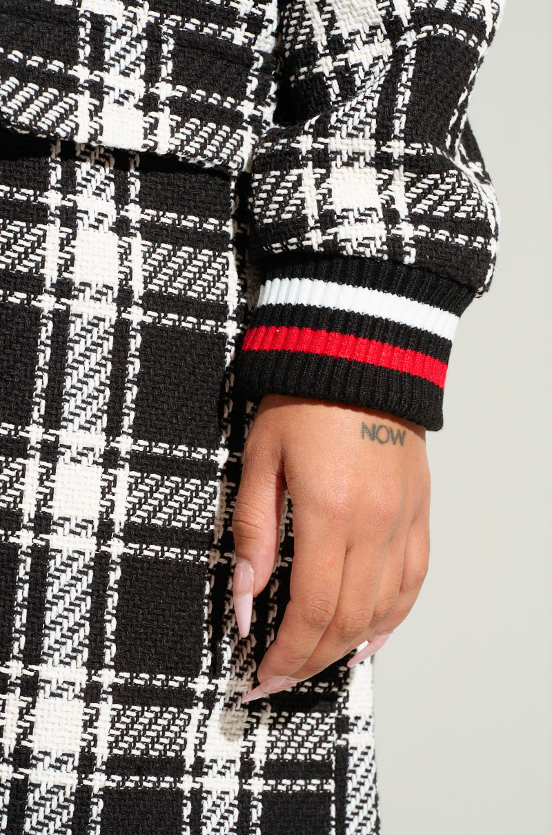 A close-up shows a person's hand resting beside their skirt, dressed in the LIBERTY VARSITY TRENCH—a black and white plaid outfit with a black cuff adorned with white and red stripes, echoing letterman-inspired patches. The person's long, manicured nails are complemented by a "NOW" tattoo on their wrist.