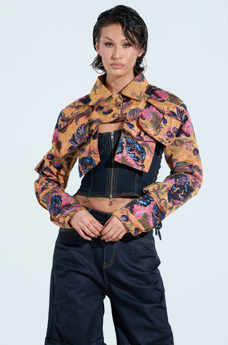 A person with short dark hair wears the ULTRA CROP CARGO BOMBER in a colorful floral pattern over a black corset top, paired with loose-fitting dark pants against a plain light blue background.