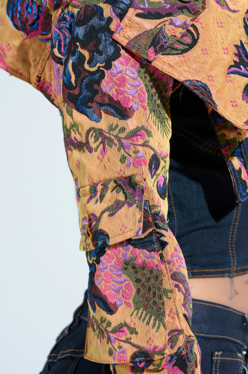 Close-up of a person wearing the ULTRA CROP CARGO BOMBER featuring colorful floral patterns in pink, purple, and green on mustard. The sleeves are rolled up to show metallic accents. The look is completed with dark denim jeans showcasing visible stitching.