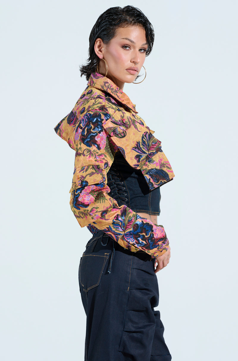 A person with short hair is wearing the ULTRA CROP CARGO BOMBER—a vibrant metallic floral jacket with ruffles—paired with dark pants. They stand in profile against a plain background, gazing forward.