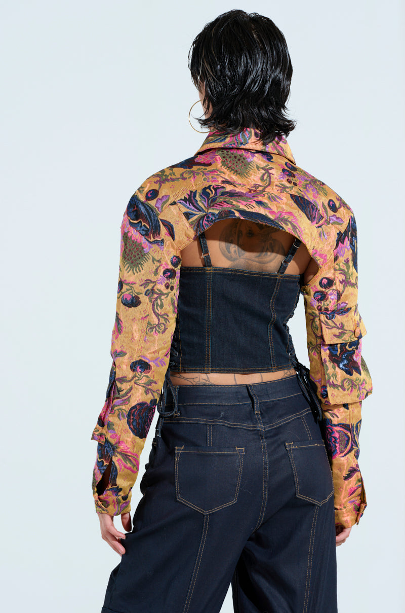 Wearing the ULTRA CROP CARGO BOMBER, a person stands facing away in a chic floral jacket with denim details and an exposed back. They pair it with dark, wide-legged pants. Their hair is styled in a short, wet look.