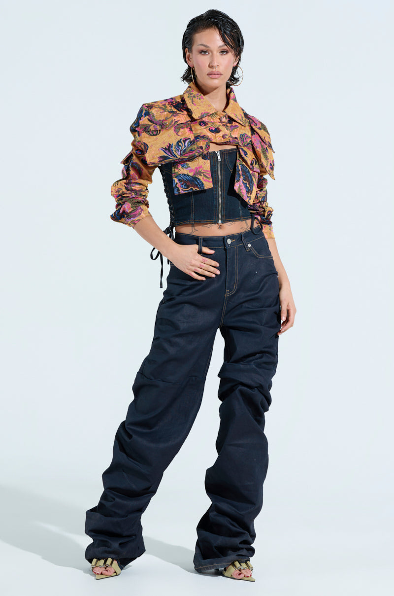 A person stands confidently wearing a colorful, abstract-patterned long-sleeve shirt under the ULTRA CROP CARGO BOMBER, paired with baggy dark jeans and open-toed shoes. The dark, structured corset adds an edge against the plain, light-colored background.