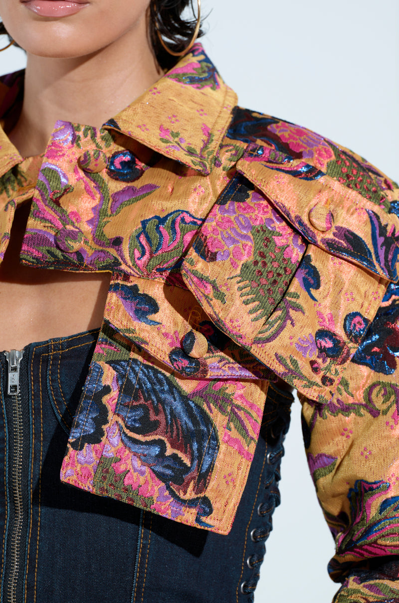 A person is seen wearing an ULTRA CROP CARGO BOMBER, a vibrant multicolored jacket with floral patterns and flap pockets in shades of yellow, pink, and blue. They're also sporting a dark denim top with visible stitching.