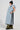 A person styled in the Fallon Tearaway Vest in Denim, featuring a long design with light blue padding and an oversized hood with black stripe accents. They complete their look with a black beanie, white shirt, green pants, and black shoes while standing in profile against a plain background.