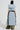 A person is wearing the Fallon Tearaway Vest in Denim, a long, sleeveless padded jacket with an oversized hood and a contrast waist belt. They complement it with a black beanie, wide-leg tie-dye pants, and chunky black boots while facing away from the camera.