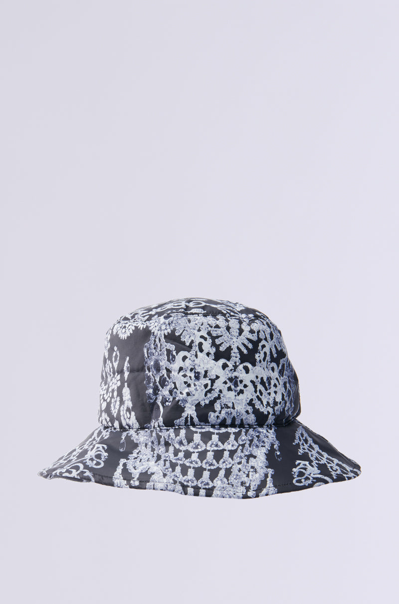 The UNDER THE LIGHTS PUFFER BUCKET HAT in black is adorned with intricate white lace-like floral patterns, elegantly sitting against a plain light gray background, reminiscent of sparkling diamonds and jewels. 1