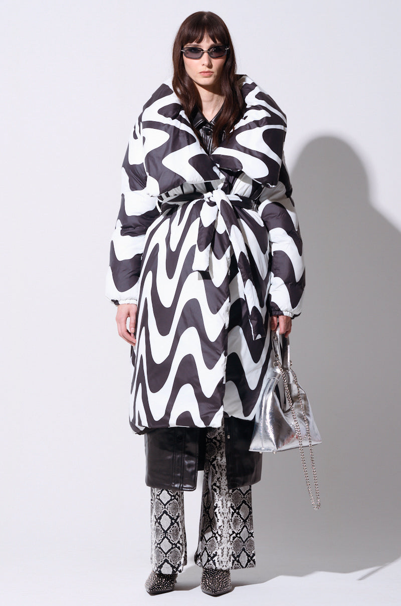 Dressed in the fashionable CARLISLE WRAP PUFFER coat, which features a striking black and white design with a bold collar and chic belt, this individual pairs it with patterned pants and accessorizes with a sleek silver handbag and sunglasses. The ensemble showcases an innovative blend of textures and prints, achieving a contemporary and stylish appearance.