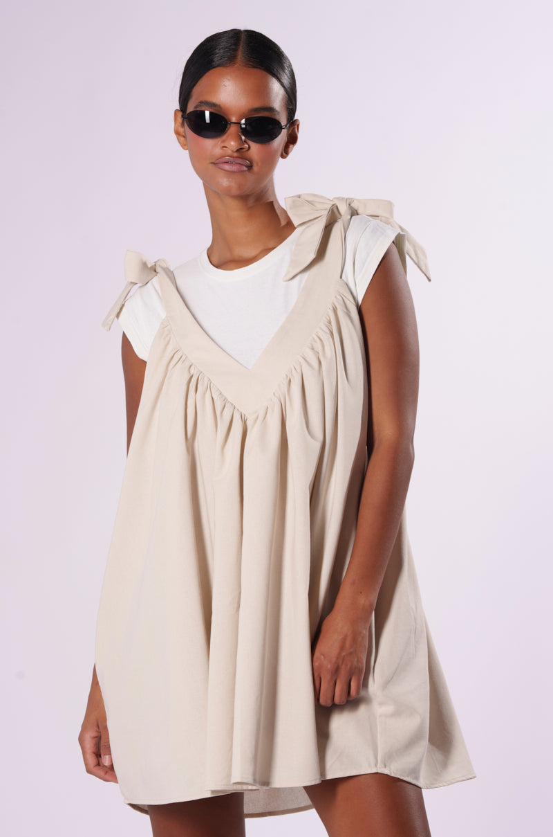 A person wearing the DONGGUAN CITY SHENGSHI ENDLESS SUMMER TIE STRAP LINEN MINI DRESS, a light, sleeveless, v-neck mini dress with bow-tied shoulder straps, layered over a white t-shirt. They have dark sunglasses and their hair is styled slicked back. The background is a light gradient.