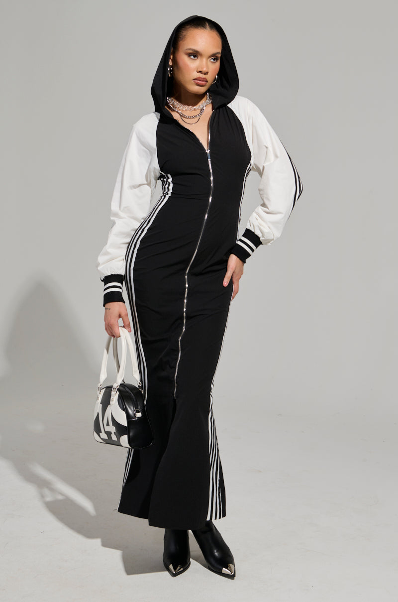 A woman confidently poses in her sporty chic ensemble, wearing the DO YOU MIND MAXI DRESS featuring a black and white color-block design with a zipper front and striped sleeves. She carries a black handbag adorned with the number 14, pairs it with heeled boots, and accessorizes with hoop earrings and necklaces.