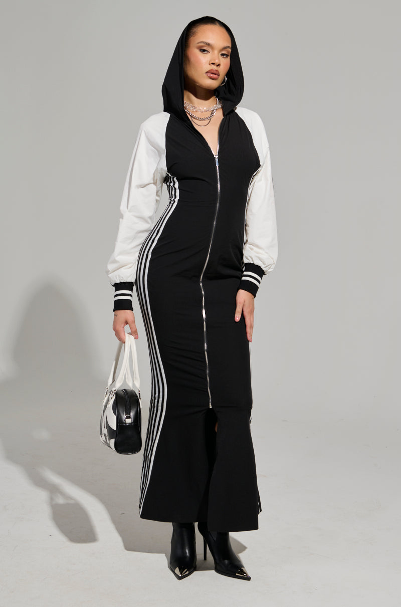 The person exudes a sporty chic vibe, dressed in the DO YOU MIND MAXI DRESS—a hooded black and white zip-up dress with striped sleeves and side details. Paired with black boots and a color-block design bag, they stand confidently against a plain background.