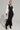 A woman poses confidently in the DO YOU MIND MAXI DRESS, a sporty chic black and white zip-up design featuring color-block panels and striped sleeves. She accessorizes with a matching handbag, black heeled boots, layered necklaces, and wears her hair styled in a sleek bun.