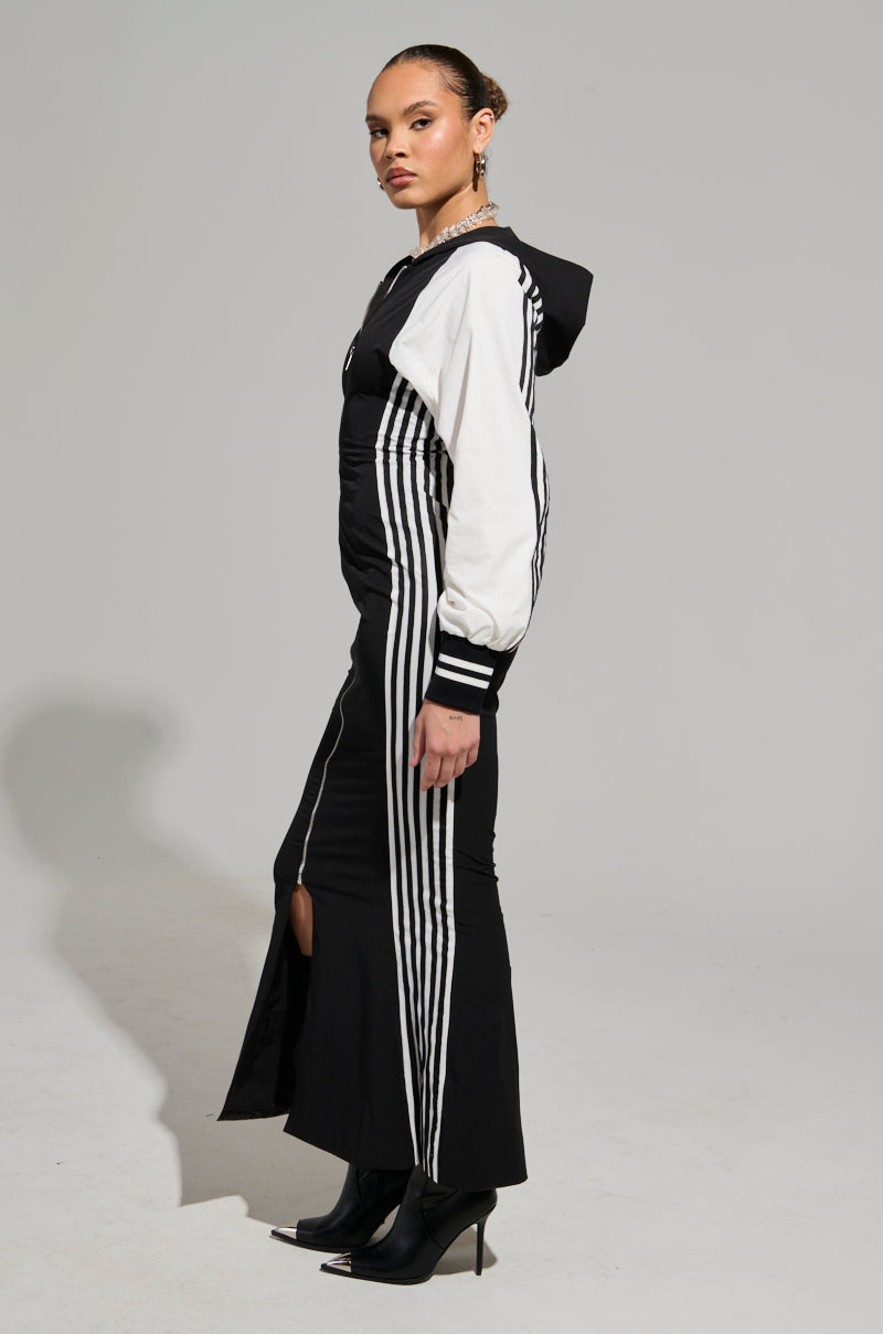 A person models the DO YOU MIND MAXI DRESS, a stylish black and white dress with side stripes, against a light gray background. The color-block design features a zippered front and a slit, perfectly paired with high-heeled ankle boots. With short hair, the person is turned to the side, highlighting their sporty chic look.