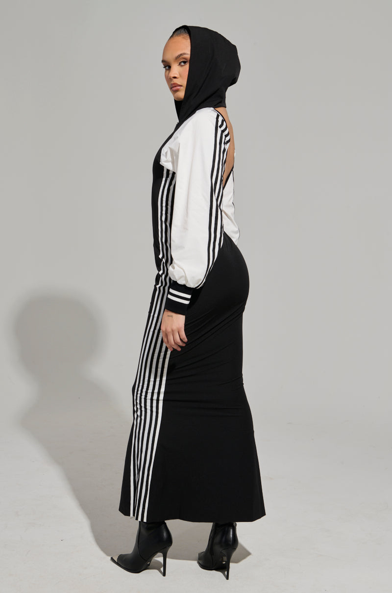 A person is dressed in the DO YOU MIND MAXI DRESS, a black and white ensemble with vertical stripes and a hood. They completed the look with black high-heeled ankle boots while posing against a plain gray background, glancing over their shoulder.