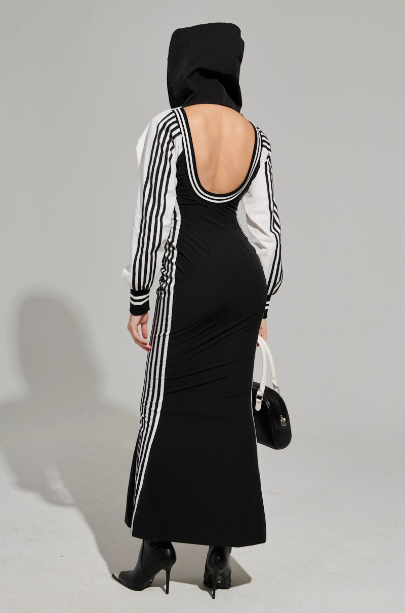 Against a plain gray background, someone exudes sporty chic vibes in the DO YOU MIND MAXI DRESS featuring a black and white color-block design with striped sleeves and an open back. They complete the look with a black hood, high-heeled boots, and a sleek black and white handbag.
