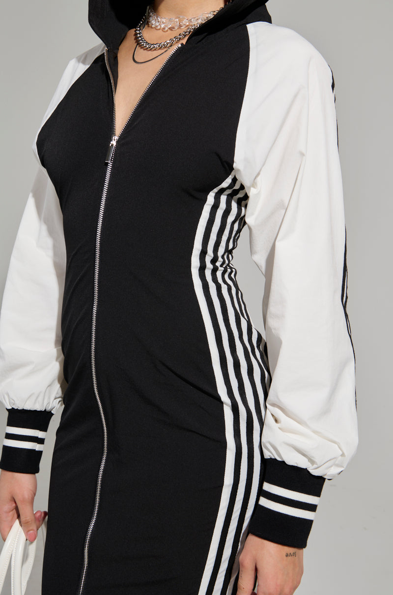 A person donning a chic DO YOU MIND MAXI DRESS, complemented by a stylish black jacket featuring a color-block design with white sleeves and side stripes. The jacket is equipped with a front zipper and sporty cuffs, capturing the essence of sporty chic flawlessly. A silver chain necklace adorns their neck for an added touch of elegance.