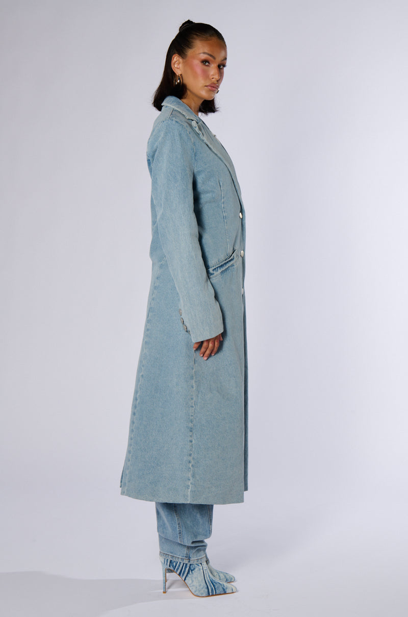 A person stands in profile against a plain background, wearing the DIVINE TIMING DENIM TRENCH over matching denim pants and high-heeled denim boots. Their hair is tied back, and they are looking slightly to the left with a neutral expression.