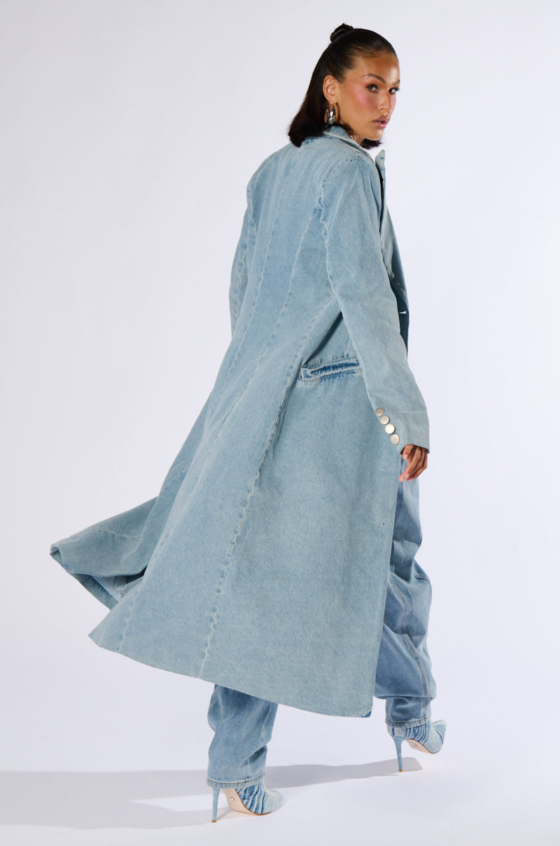 A person with long, dark hair styled in a high ponytail is wearing the DIVINE TIMING DENIM TRENCH paired with matching wide-legged denim pants and pointed high heels. As they look back over their shoulder, the trench coat flares out slightly with their movement.