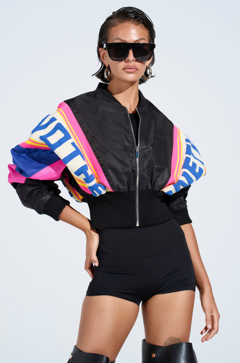 Wearing the NEW PHOENIX MIXED MEDIA BOMBER, a person pairs it with black high-waisted shorts, knee-high boots, and large sunglasses. Their slicked-back hair perfects the bold look as they confidently pose against a plain backdrop.