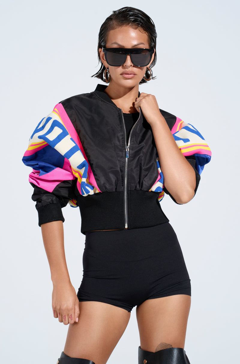 Wearing oversized black sunglasses and a NEW PHOENIX MIXED MEDIA BOMBER with puffy sleeves in a vibrant colorblock design, they pair it with black high-waisted shorts and knee-high boots while posing confidently against a plain background.