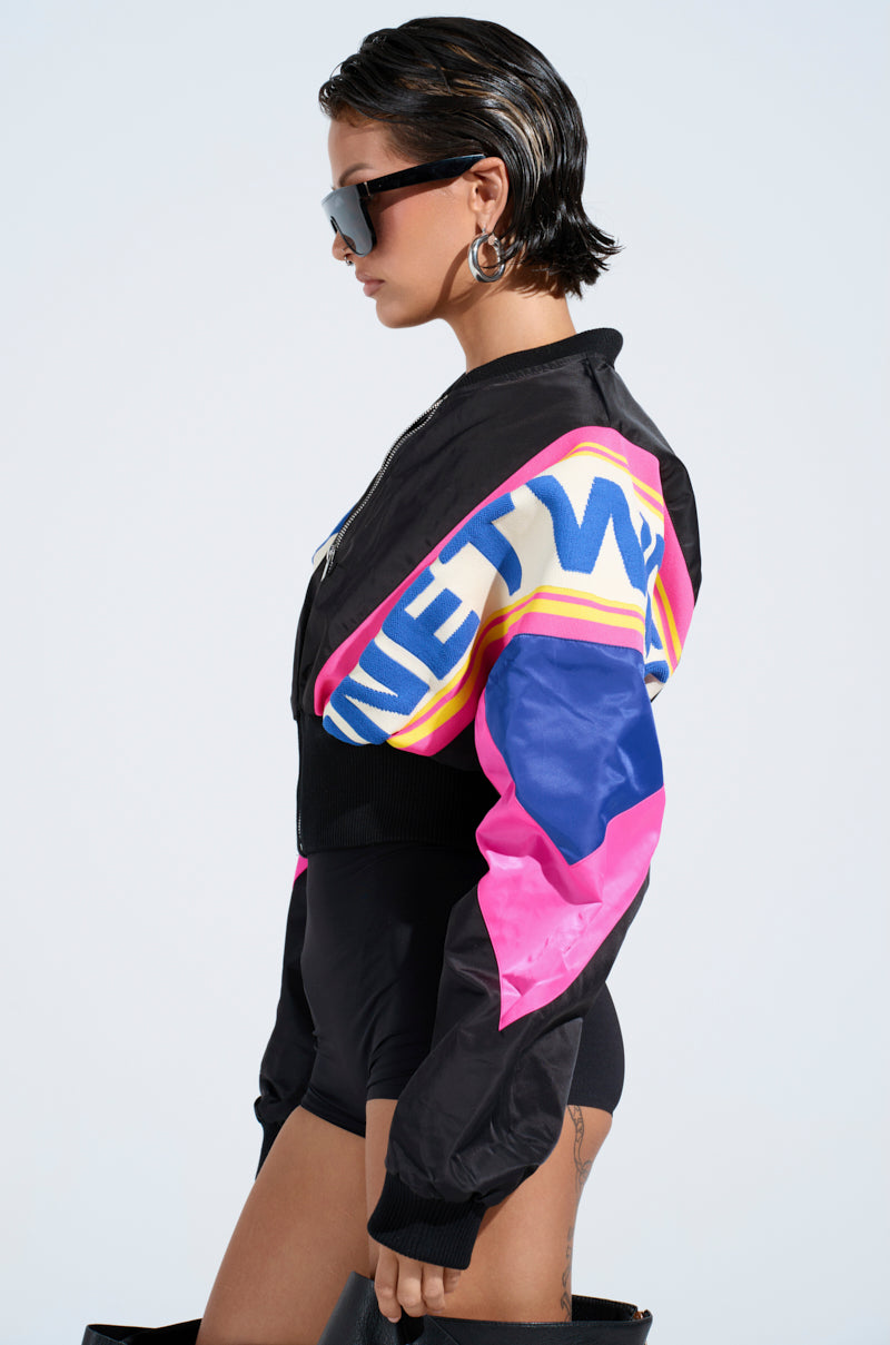 With short hair and sunglasses, a person embodies retro chic in the NEW PHOENIX MIXED MEDIA BOMBER featuring bold sleeve lettering. They effortlessly pair it with dark shorts and knee-high boots, posing in profile against a plain background.