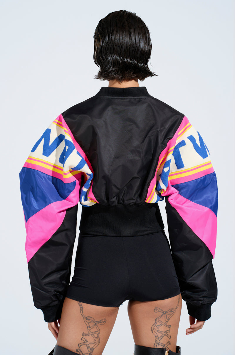 Wearing the vibrant NEW PHOENIX MIXED MEDIA BOMBER with pink, blue, and yellow graphics, a person stands with their back to the camera. Their short dark hair complements black shorts and thigh-high boots, revealing striking leg tattoos.
