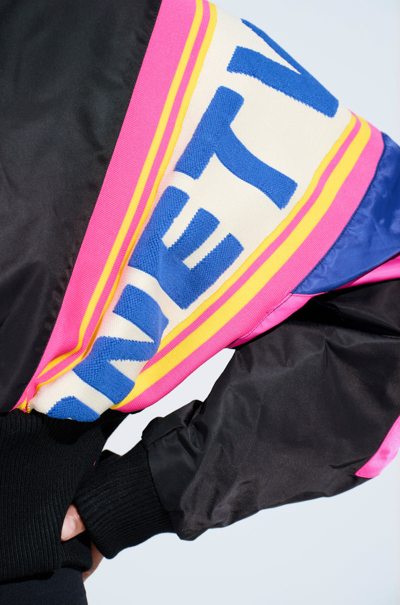 A close-up showcases a person wearing the vibrant NEW PHOENIX MIXED MEDIA BOMBER. The sleeve features a colorblock design with blue lettering, yellow and pink stripes against black fabric. The person's hands are tucked in at the waist.