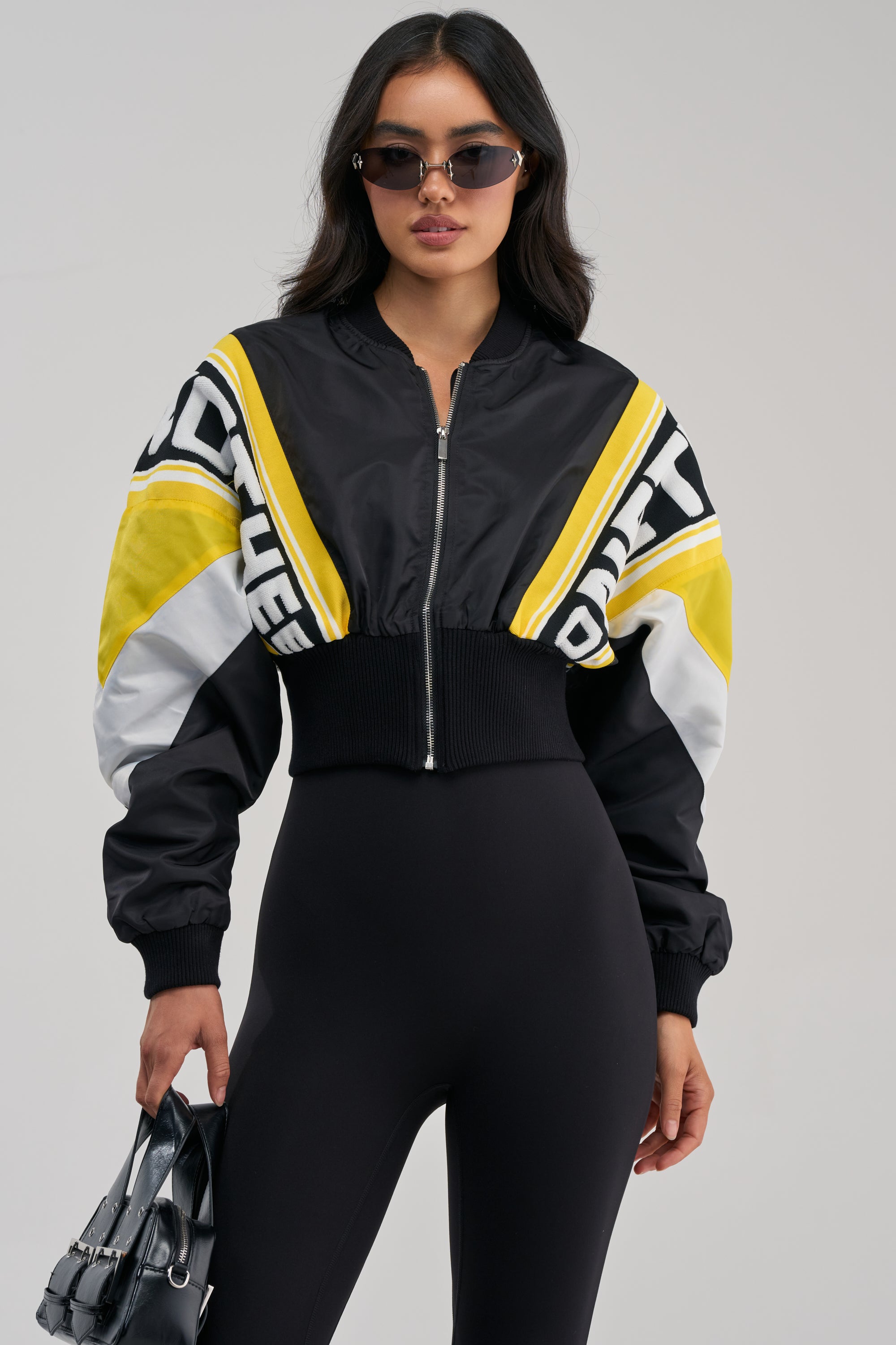 A woman in sunglasses wears the PHOENIX MIXED MEDIA BOMBER IN YELLOW—a black and yellow streetwear jacket with white accents—layered over a fitted black outfit. She holds a small purse in her left hand, standing against a plain light background.