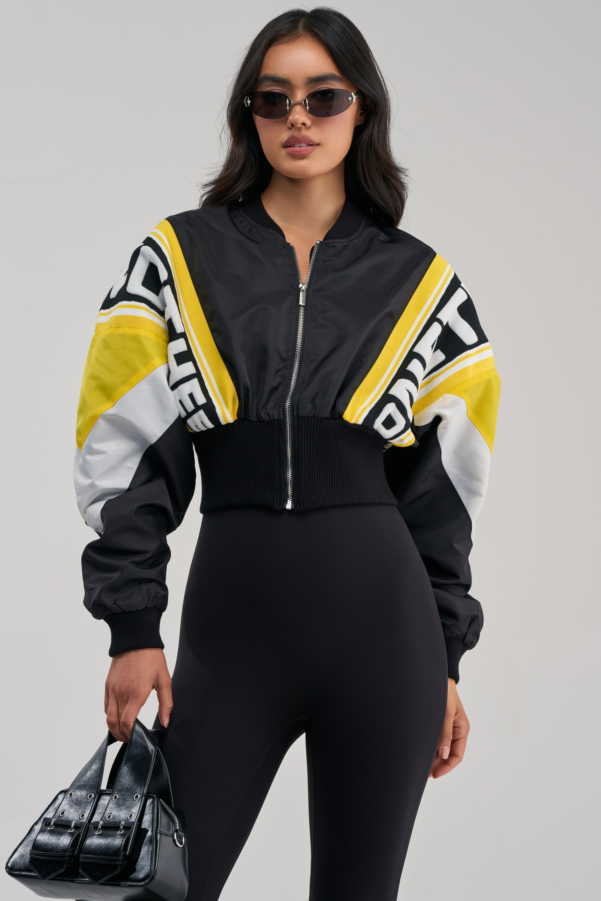 A woman wearing sunglasses, the PHOENIX MIXED MEDIA BOMBER IN YELLOW, and a black jumpsuit holds a shiny black handbag against a plain background.