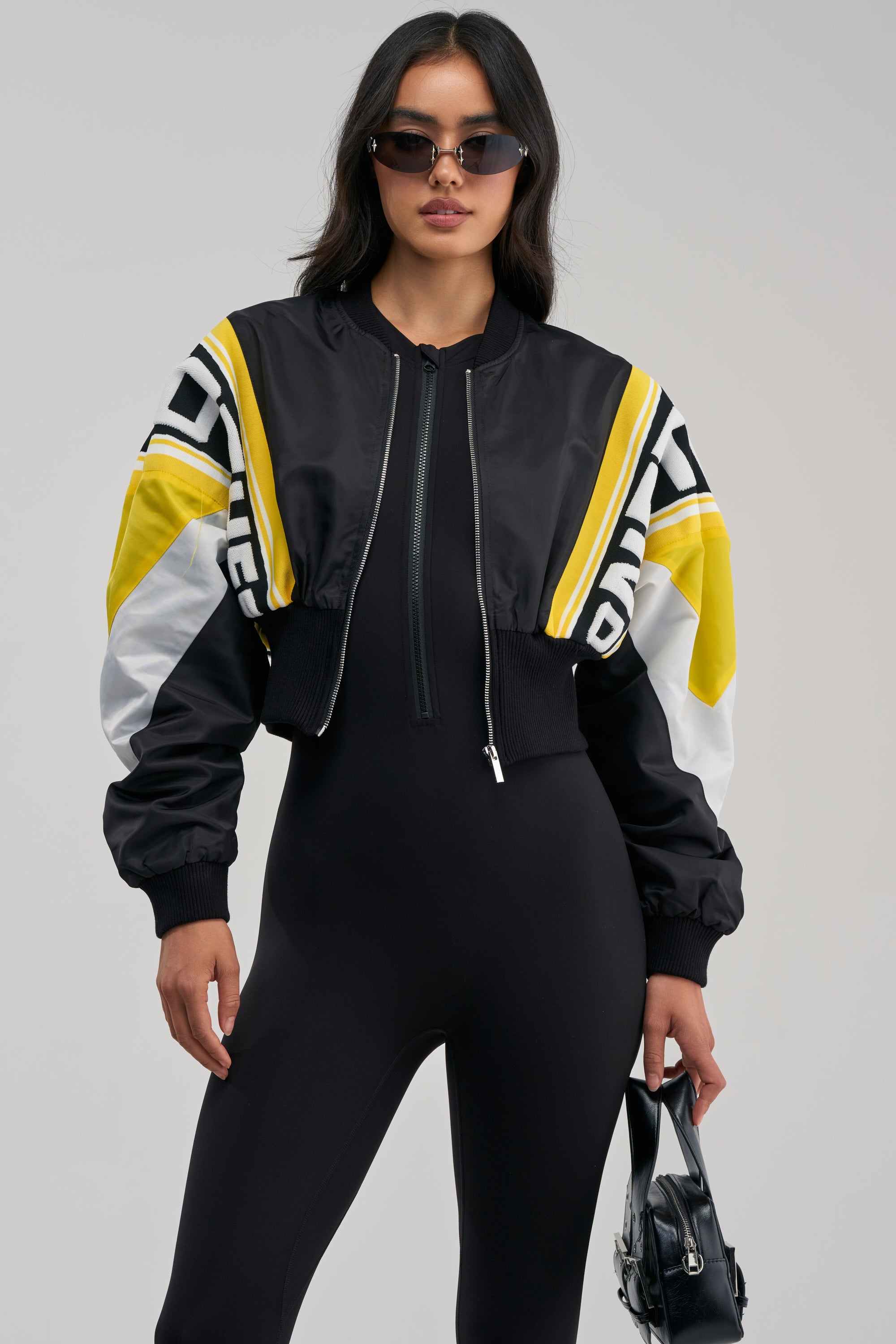 A woman in black sunglasses and a black jumpsuit wears the PHOENIX MIXED MEDIA BOMBER IN YELLOW, standing against a plain light background and holding a shiny black handbag.