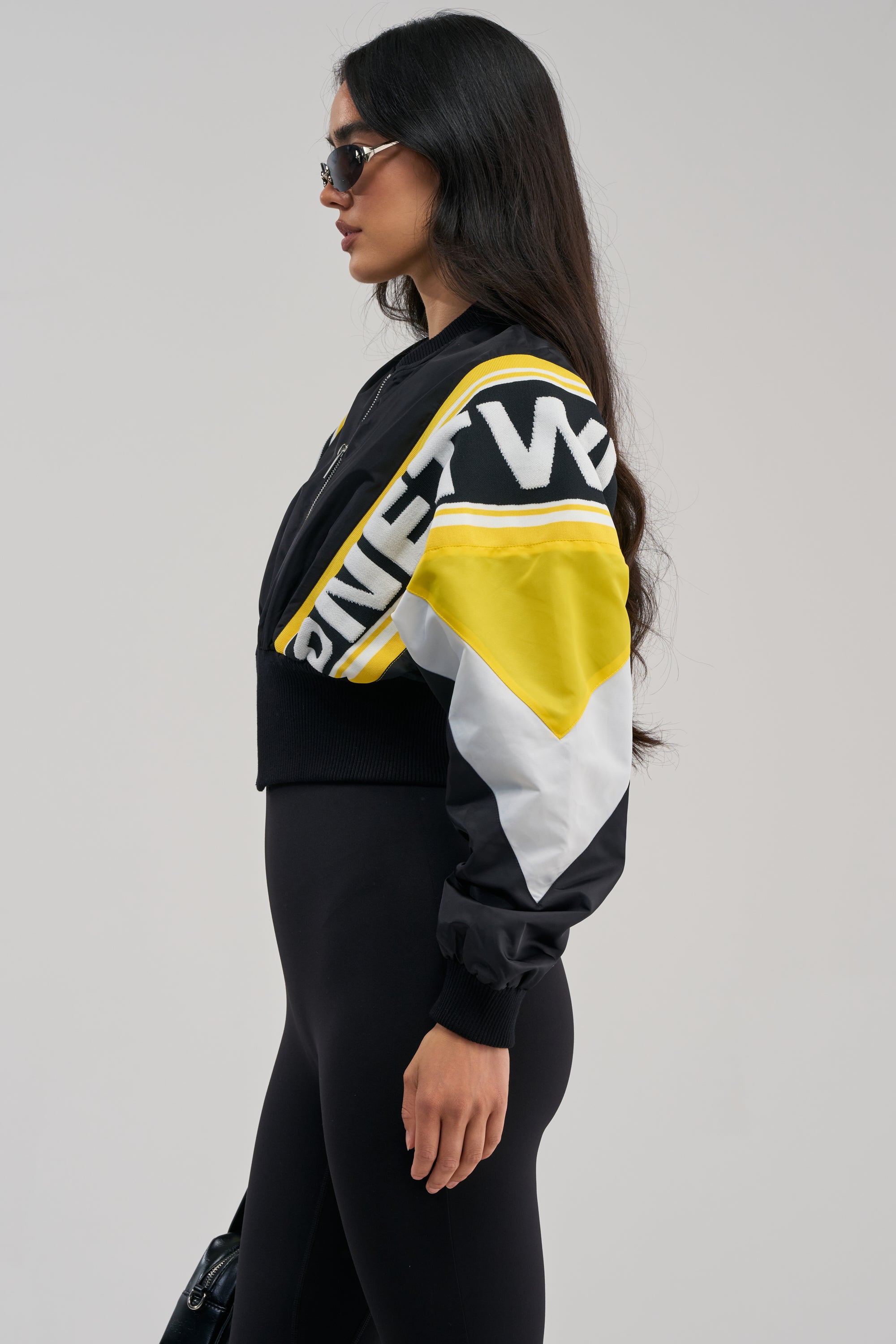 Woman in profile wearing black sunglasses, the PHOENIX MIXED MEDIA BOMBER IN YELLOW, and black leggings, standing against a plain background.