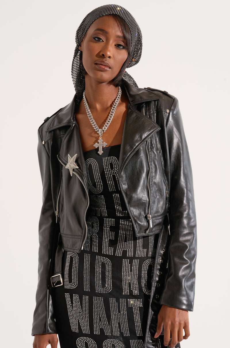 A woman poses in the Smooth Operator Faux Lamb Skin Moto jacket, a cropped silhouette adorned with a star pin. She wears it over a black dress with white text and accessorizes with a sparkling head scarf and a large chain necklace featuring a cross pendant. Silver metallic hardware on the jacket adds an edgy touch to her serious expression and partially covered hair. 2