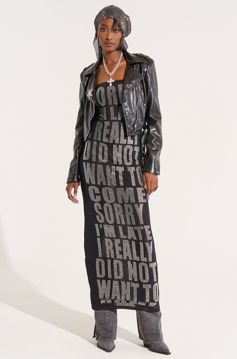 A woman stands confidently wearing the Smooth Operator Faux Lambskin Moto jacket with silver metallic hardware over a long black dress adorned with silver text, a sparkling headwrap, a long necklace, and matching grey boots. The text on the dress reads various phrases in silver, including "SORRY I'M LATE. 5