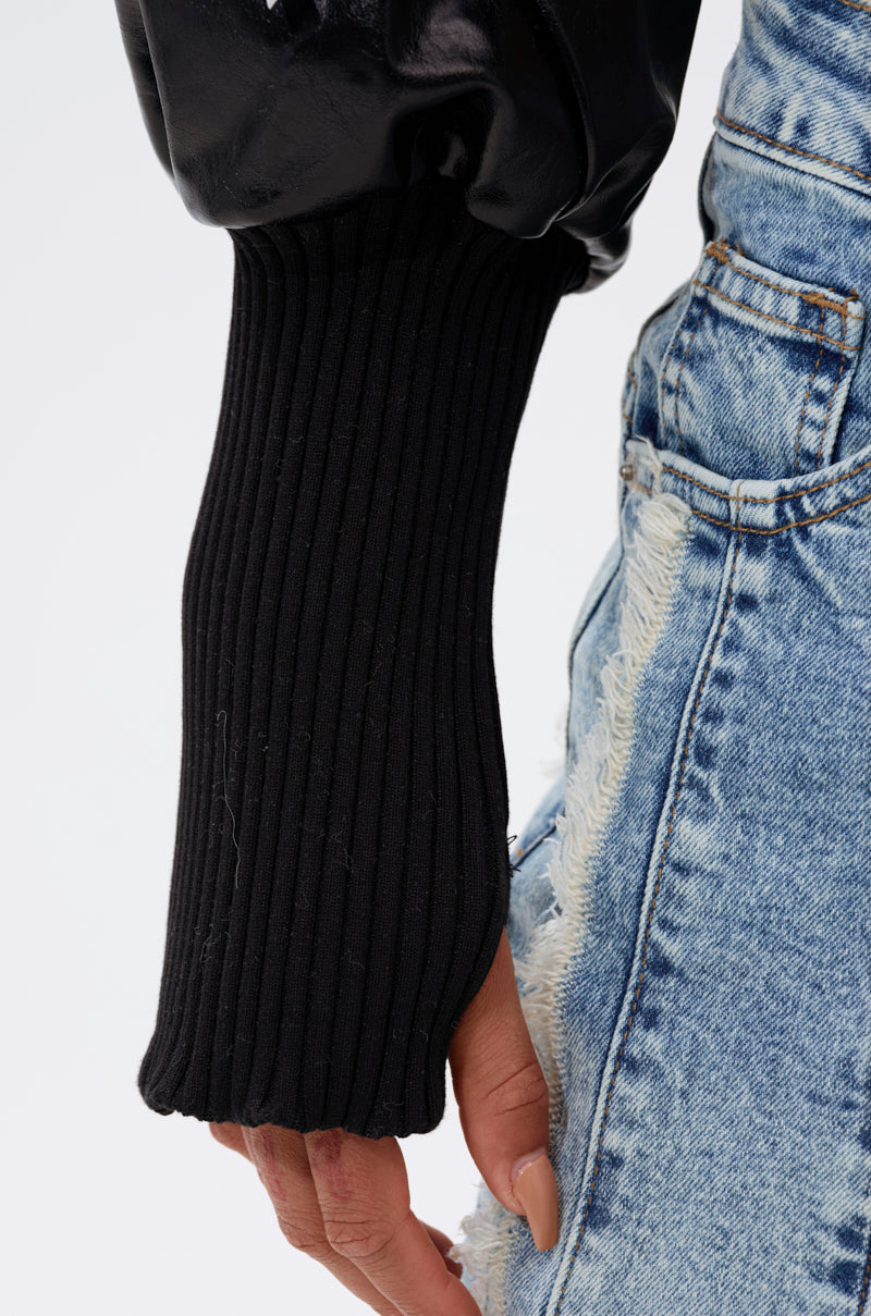 A close-up of a person wearing the NOW YOU SEE ME BOLERO, featuring black ribbed sleeves with thumb holes, styled with distressed blue denim jeans. The hand rests alongside the hip, partially visible. 10