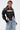 A woman with wet, slicked-back hair wears the NOW YOU SEE ME BOLERO over a black denim corset top with white accents and high-waisted distressed blue jeans, confidently posing against a plain background.