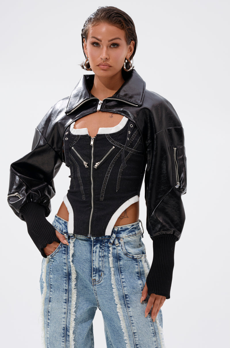 A woman wears the NOW YOU SEE ME BOLERO, a cropped jacket with oversized sleeves, styled over a zippered corset top and two-tone patchwork jeans. With wet-look hair and hoops, it's perfect for transitional outerwear style. 2