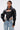 A woman wears the NOW YOU SEE ME BOLERO, a cropped jacket with oversized sleeves, styled over a zippered corset top and two-tone patchwork jeans. With wet-look hair and hoops, it's perfect for transitional outerwear style.