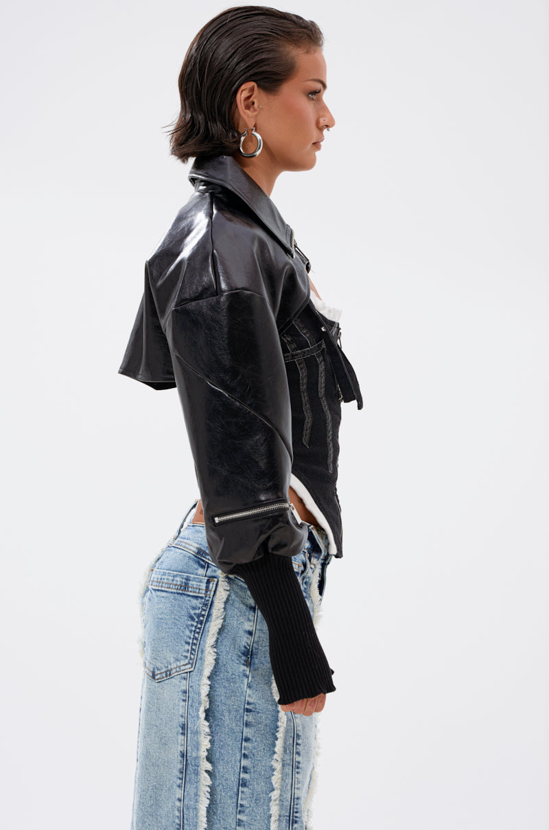 A woman with short, slicked-back hair models the NOW YOU SEE ME BOLERO, a shiny black faux leather piece with long sleeves, styled with distressed light-wash jeans. She stands in profile against a plain white background. 4