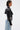 A woman with short, slicked-back hair models the NOW YOU SEE ME BOLERO, a shiny black faux leather piece with long sleeves, styled with distressed light-wash jeans. She stands in profile against a plain white background.