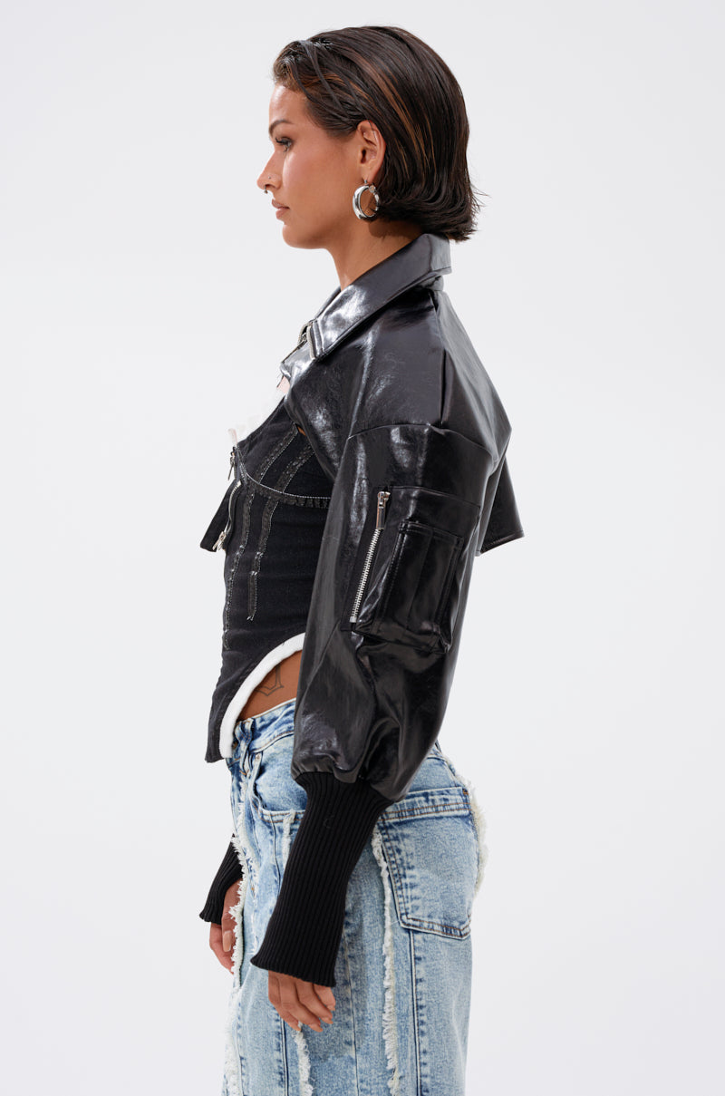 A woman with short dark hair stands in profile, wearing the NOW YOU SEE ME BOLERO in glossy black faux leather with ribbed cuffs and light blue distressed jeans. She accessorizes with hoop earrings, her hands resting by her sides. 5