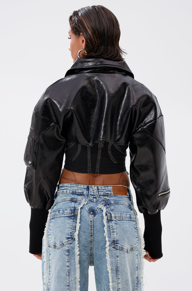 Someone with short dark hair wears the NOW YOU SEE ME BOLERO, a shiny black faux leather bolero with puffy sleeves and ribbed cuffs, paired with high-waisted light blue distressed jeans, standing facing away from the camera. 6