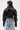 Someone with short dark hair wears the NOW YOU SEE ME BOLERO, a shiny black faux leather bolero with puffy sleeves and ribbed cuffs, paired with high-waisted light blue distressed jeans, standing facing away from the camera.