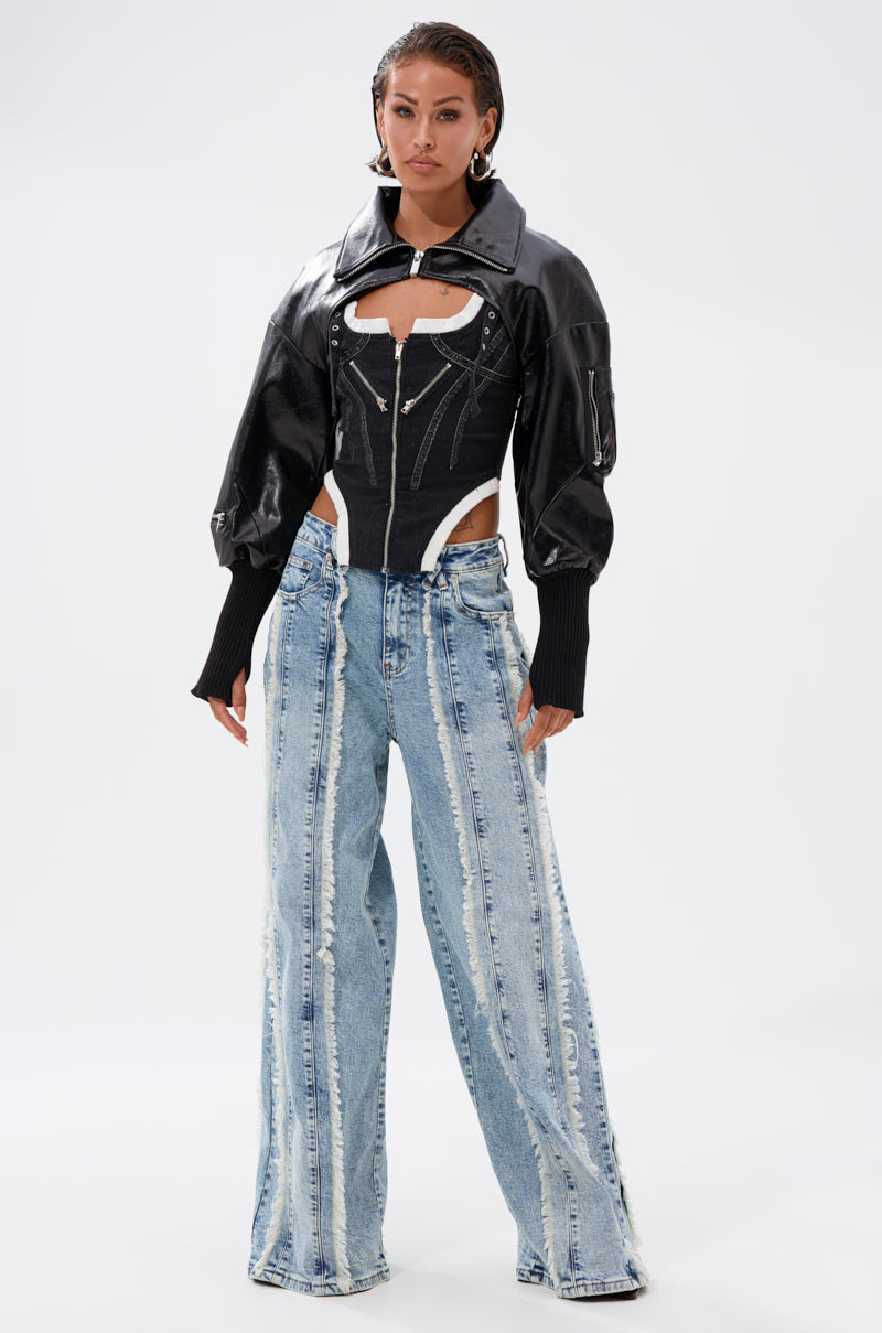 A woman stands confidently in the NOW YOU SEE ME BOLERO—a black faux leather jacket with puffy sleeves—paired with a black corset-style top and wide-leg light blue distressed jeans with vertical seam details. 7
