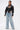A woman stands confidently in the NOW YOU SEE ME BOLERO—a black faux leather jacket with puffy sleeves—paired with a black corset-style top and wide-leg light blue distressed jeans with vertical seam details.