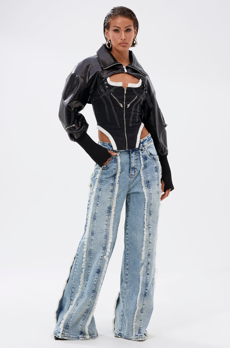 A woman poses in the NOW YOU SEE ME BOLERO, styled with a black cutout corset top and high-waisted wide-leg jeans with frayed seams. She accessorizes with large hoop earrings and slicked-back hair for a chic, transitional look. 8
