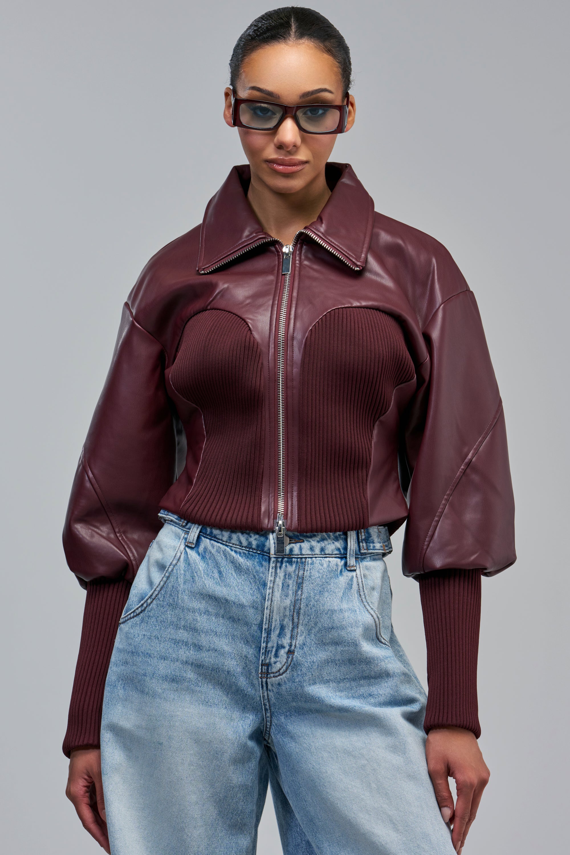 A person wears the BETTER LUCK NEXT TIME FITTED BOMBER IN BURGUNDY, a faux leather moto jacket with a zippered front, ribbed panels, and puffy sleeves, paired with light-wash jeans and dark-rimmed glasses.