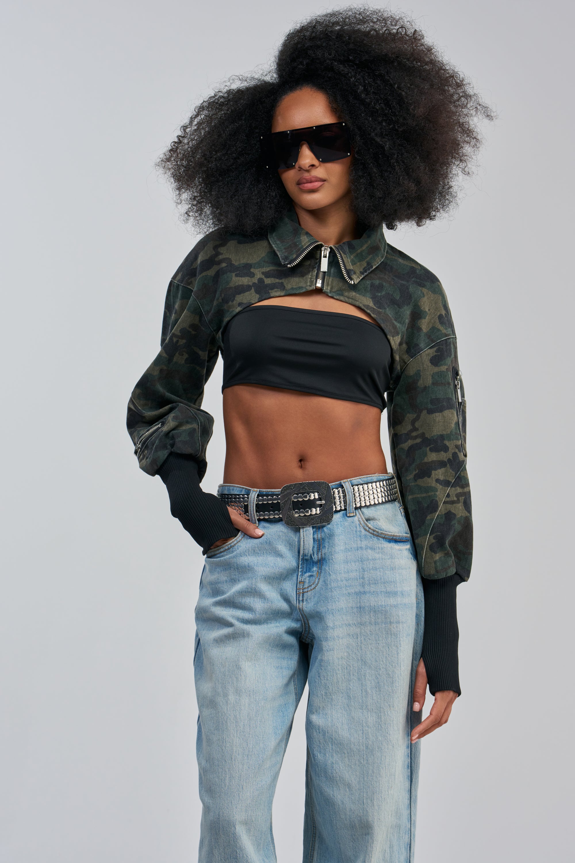 A woman with voluminous curly hair and large sunglasses poses confidently against a plain background, layering the NOW YOU SEE ME CAMO BOLERO in true streetwear fashion over a black tube top, styled with a studded belt and blue jeans.